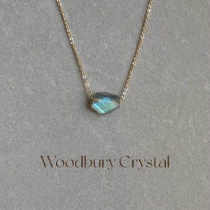 May include: A delicate gold chain necklace with a single, faceted, iridescent blue-green labradorite gemstone pendant. The pendant is a simple, geometric shape and hangs from a delicate chain. The necklace is labeled "Woodbury Crystal".