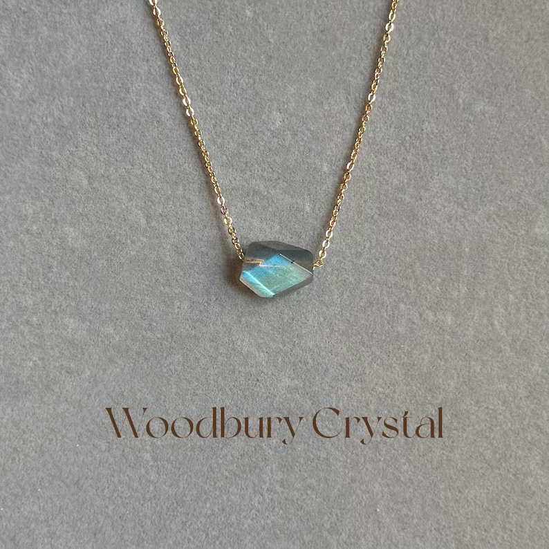 May include: A delicate gold chain necklace with a single, faceted, iridescent blue-green labradorite gemstone pendant. The pendant is a simple, geometric shape and hangs from a delicate chain. The necklace is labeled "Woodbury Crystal".