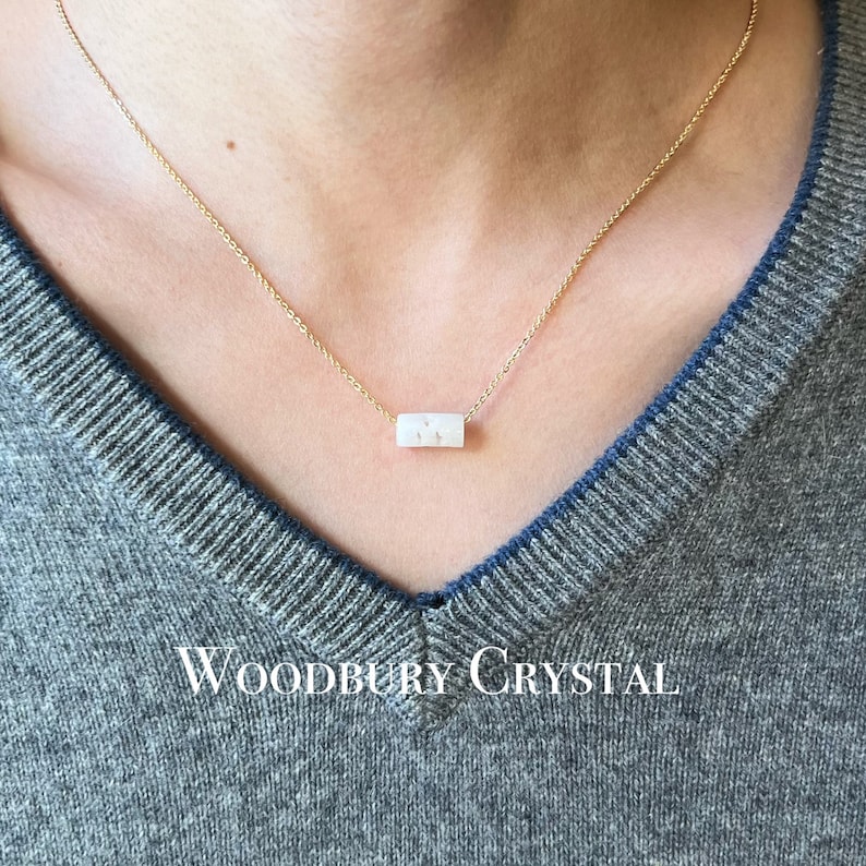 May include: A delicate gold chain necklace with a white, rectangular pendant. The pendant has a subtle sparkle and the text "Woodbury Crystal" is visible at the bottom of the image.