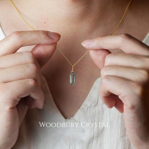 May include: A gold chain necklace with a light green crystal pendant in the shape of a long, thin, pointed rectangle. The text "WOODBURY CRYSTAL" is at the bottom of the image.
