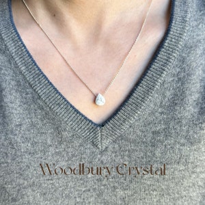 May include: A silver chain necklace with a white teardrop-shaped pendant. The pendant has a marbled pattern. The necklace is worn by a person wearing a gray sweater. The text "Woodbury Crystal" is visible at the bottom of the image.