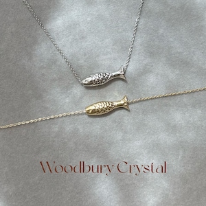 May include: Two delicate necklaces with fish pendants. One necklace has a silver fish pendant, and the other has a gold fish pendant. The necklaces are on a light brown surface with the text "Woodbury Crystal" below the necklaces.