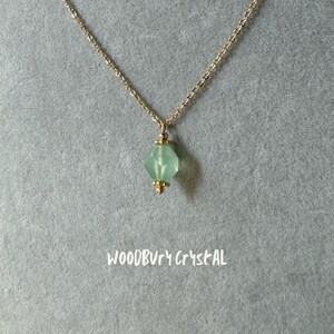Op de afbeelding: Een delicate gouden ketting met een lichtgroene gefacetteerde kristallen hanger. De hanger heeft een gouden zetting en is versierd met kleine gouden kralen. De ketting wordt getoond op een grijze achtergrond met de tekst "WOODBURY CRYSTAL" onder de hanger.