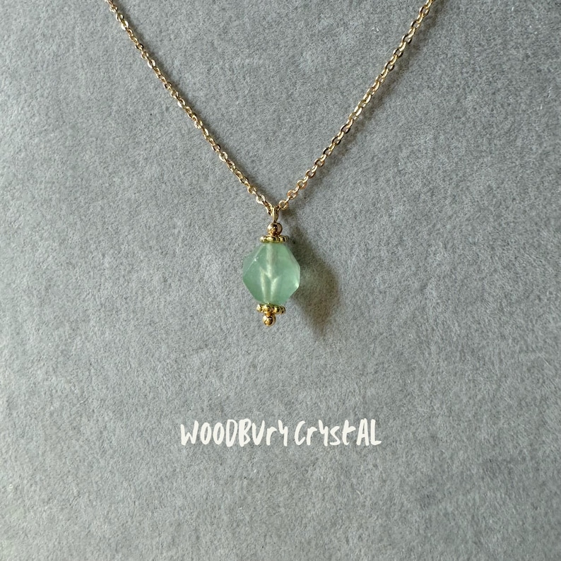 Op de afbeelding: Een delicate gouden ketting met een lichtgroene gefacetteerde kristallen hanger. De hanger heeft een gouden zetting en is versierd met kleine gouden kralen. De ketting wordt getoond op een grijze achtergrond met de tekst "WOODBURY CRYSTAL" onder de hanger.
