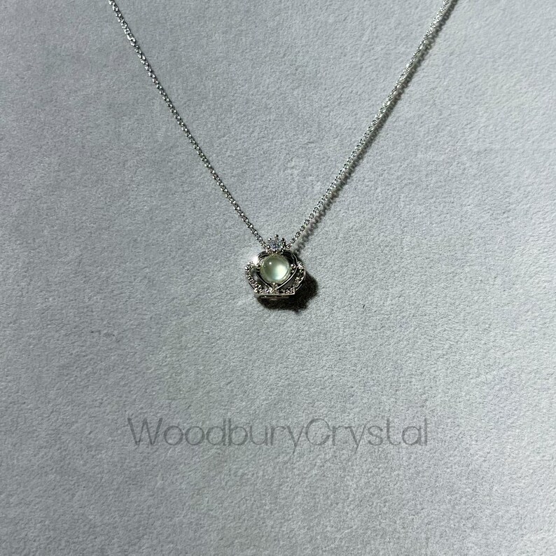 Puede incluir: Un collar de plata con un colgante en forma de coraz&oacute;n que presenta una piedra preciosa verde claro rodeada de piedras preciosas transparentes m&aacute;s peque&ntilde;as. El colgante est&aacute; engastado en un dise&ntilde;o de corona.