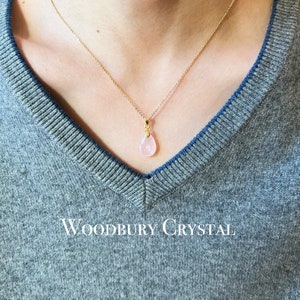 May include: A delicate gold chain necklace with a pink teardrop-shaped pendant. The pendant is made of rose quartz and is set in a gold setting. The necklace is on a model wearing a gray sweater. The text "WOODBURY CRYSTAL" is visible at the bottom of the image.