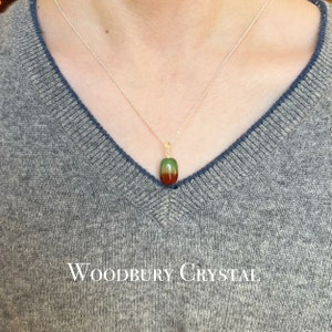 May include: A silver chain necklace with a green, red, and brown oval pendant. The pendant is made of a natural stone. The necklace is on a person wearing a gray sweater. The text "WOODBURY CRYSTAL" is visible at the bottom of the image.