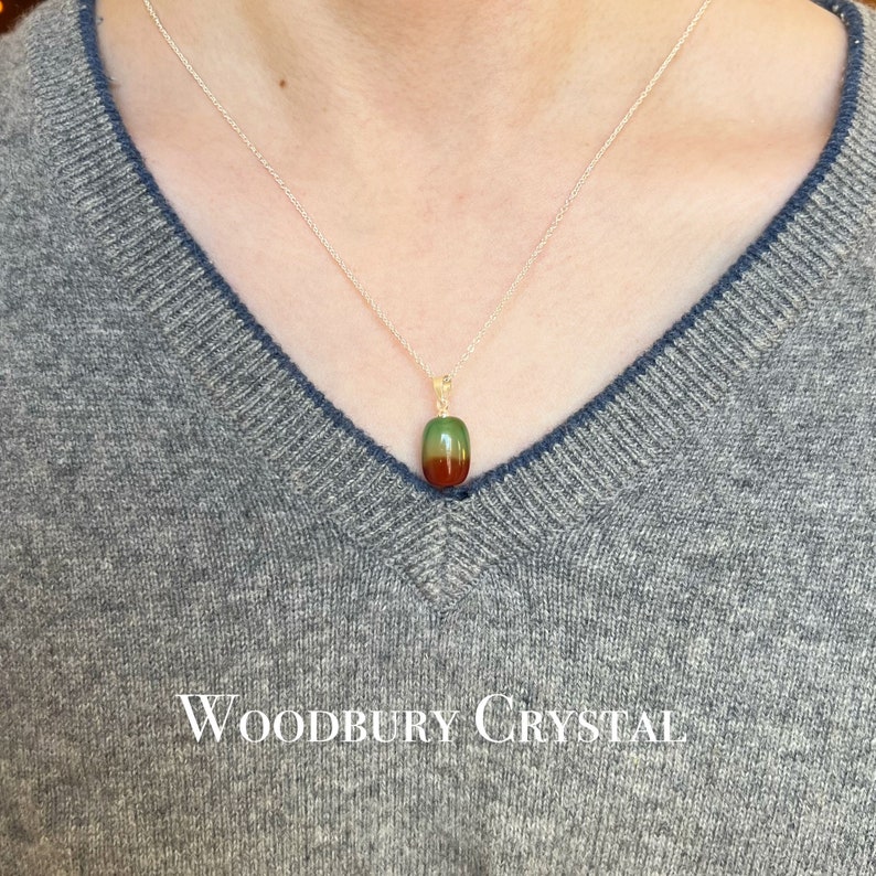 May include: A silver chain necklace with a green, red, and brown oval pendant. The pendant is made of a natural stone. The necklace is on a person wearing a gray sweater. The text "WOODBURY CRYSTAL" is visible at the bottom of the image.