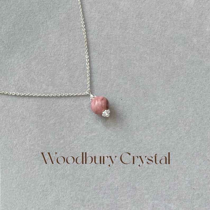 May include: A silver chain necklace with a pink and white gemstone pendant. The pendant is a round, smooth stone with a silver setting. The text "Woodbury Crystal" is printed below the necklace.