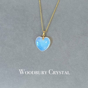 May include: A gold chain necklace with a light blue heart-shaped pendant. The pendant has a iridescent finish. The necklace is on a gray background with the text "WOODBURY CRYSTAL" below the pendant.