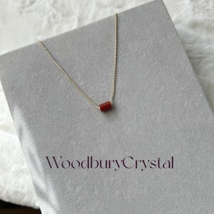 May include: A delicate gold chain necklace with a small, red, cylindrical pendant. The necklace is displayed on a gray surface with the text "Woodbury Crystal" printed below.