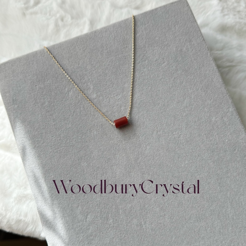 May include: A delicate gold chain necklace with a small, red, cylindrical pendant. The necklace is displayed on a gray surface with the text "Woodbury Crystal" printed below.