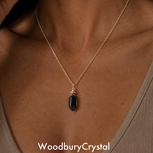 May include: A gold chain necklace featuring a single, faceted, hexagonal black crystal pendant. The pendant is secured by a small gold cap at the top. The text WoodburyCrystal is printed in white at the bottom of the image against a neutral background.