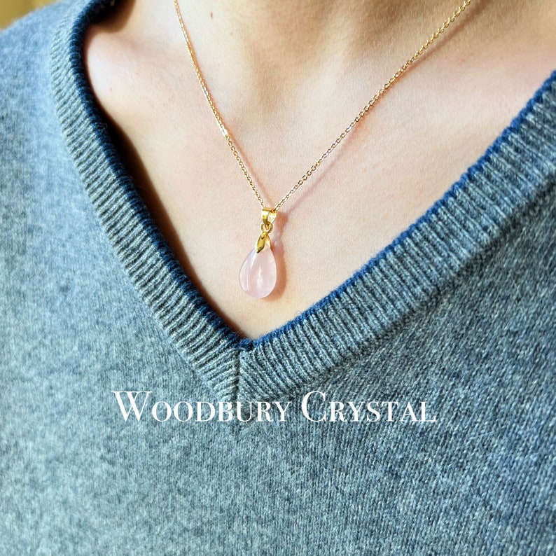 May include: A delicate gold chain necklace with a pink teardrop-shaped pendant. The pendant is made of rose quartz and is set in a gold setting. The necklace is worn by a person with a gray sweater. The text "WOODBURY CRYSTAL" is visible at the bottom of the image.