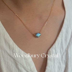 May include: A gold chain necklace with a faceted iridescent blue labradorite gemstone pendant. The necklace is on a model wearing a white shirt. The text "Woodbury Crystal" is visible at the bottom of the image.