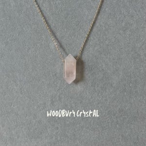 Könnte beinhalten: Eine silberne Kettenkette mit einem hellrosa Kristallanhänger in Form eines langen, dünnen, spitz zulaufenden Sechsecks. Der Anhänger hängt an der Kette. Der Text "WOODBURY CRYSTAL" ist in Weiß auf grauem Hintergrund geschrieben.