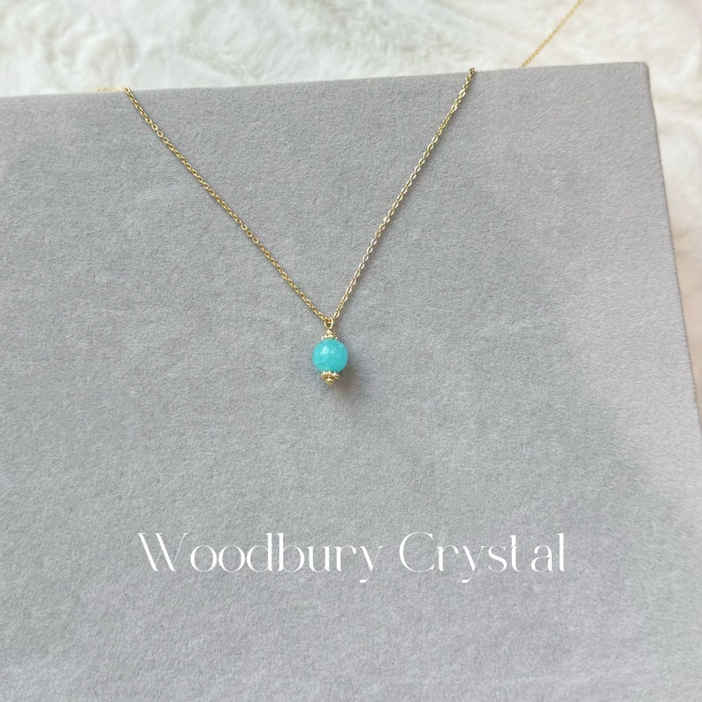 May include: A delicate gold chain necklace with a single, round turquoise gemstone pendant. The necklace is displayed on a gray background with the text "Woodbury Crystal" printed below.