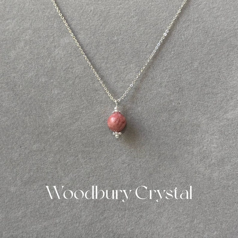 May include: A silver chain necklace with a pink and white round gemstone pendant. The pendant is accented with small silver beads. The text "Woodbury Crystal" is printed below the necklace.