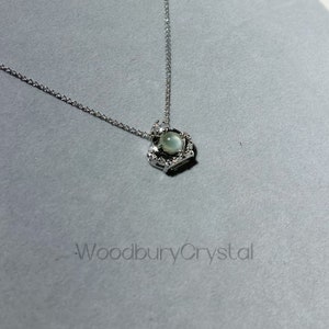 Puede incluir: Un collar de plata con un colgante en forma de coraz&oacute;n que presenta una piedra preciosa redonda de color verde claro rodeada de piedras preciosas m&aacute;s peque&ntilde;as y transparentes.