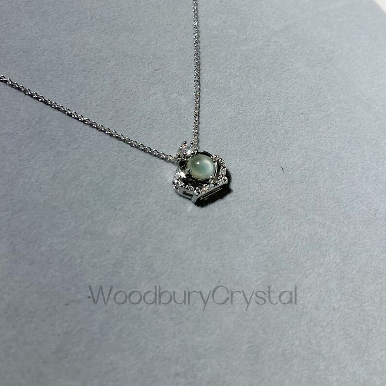 Puede incluir: Un collar de plata con un colgante en forma de coraz&oacute;n que presenta una piedra preciosa redonda de color verde claro rodeada de piedras preciosas m&aacute;s peque&ntilde;as y transparentes.