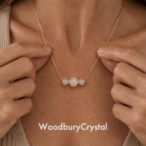 May include: A gold necklace with three pale pink gemstone beads. The necklace is shown against a neutral background. The brand name "WoodburyCrystal" is displayed at the bottom of the image.