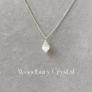 May include: A silver chain necklace with a white, round, crystal pendant. The pendant is surrounded by small silver beads. The text "Woodbury Crystal" is printed below the pendant.