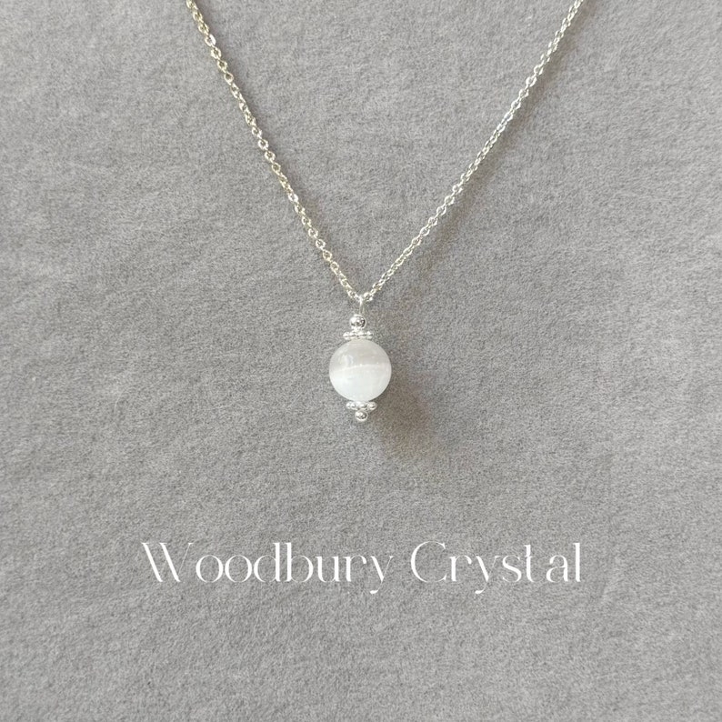May include: A silver chain necklace with a white, round, crystal pendant. The pendant is surrounded by small silver beads. The text "Woodbury Crystal" is printed below the pendant.