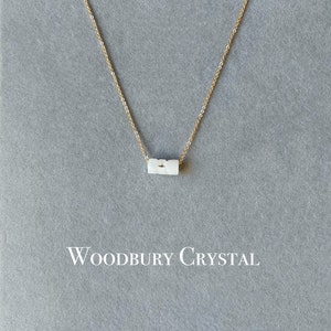 May include: A delicate gold chain necklace with a white, rectangular pendant. The pendant has a subtle, textured surface. The necklace is displayed on a gray background with the text "Woodbury Crystal" printed below.