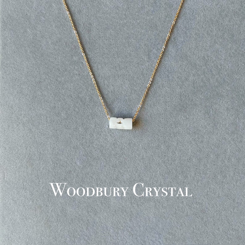 May include: A delicate gold chain necklace with a white, rectangular pendant. The pendant has a subtle, textured surface. The necklace is displayed on a gray background with the text "Woodbury Crystal" printed below.