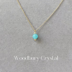 May include: A gold chain necklace with a light blue round pendant. The pendant has a gold trim. The necklace is on a gray background. The text "Woodbury Crystal" is at the bottom of the image.