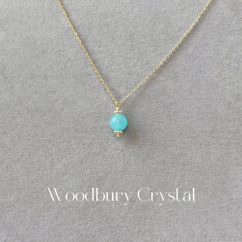 May include: A gold chain necklace with a light blue round pendant. The pendant has a gold trim. The necklace is on a gray background. The text "Woodbury Crystal" is at the bottom of the image.