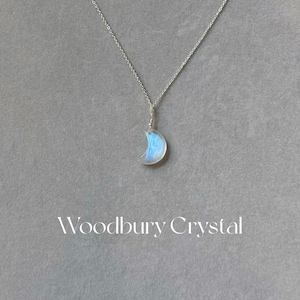 May include: A silver chain necklace with a crescent moon pendant made of iridescent blue moonstone. The pendant is about 1 inch in diameter. The text "Woodbury Crystal" is printed below the pendant.