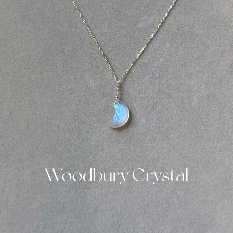 May include: A silver chain necklace with a crescent moon pendant made of iridescent blue moonstone. The pendant is about 1 inch in diameter. The text "Woodbury Crystal" is printed below the pendant.
