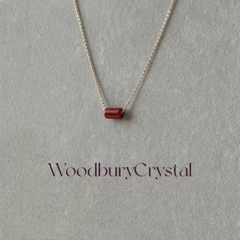 May include: A gold chain necklace with a single, red, rectangular gemstone pendant. The pendant is a simple, minimalist design. The text "WoodburyCrystal" is visible at the bottom of the image.