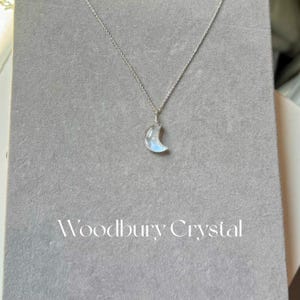 May include: A silver chain necklace with a crescent moon-shaped rainbow moonstone pendant. The pendant has a light blue iridescent sheen. The necklace is displayed on a gray background with the text "Woodbury Crystal" printed below the pendant.