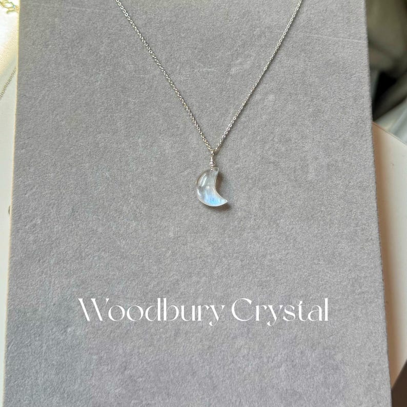 May include: A silver chain necklace with a crescent moon-shaped rainbow moonstone pendant. The pendant has a light blue iridescent sheen. The necklace is displayed on a gray background with the text "Woodbury Crystal" printed below the pendant.