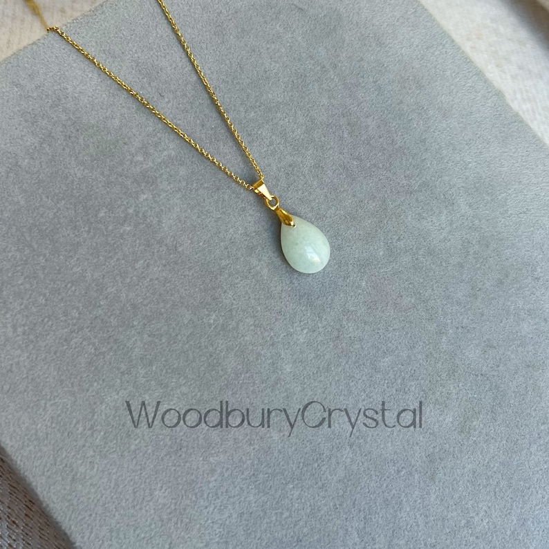Puede incluir: Un delicado collar de cadena dorada con un colgante en forma de l&aacute;grima de color verde claro. El colgante est&aacute; hecho de una piedra lisa y pulida. El collar se muestra sobre un fondo gris con el texto "WoodburyCrystal" impreso en la parte inferior.