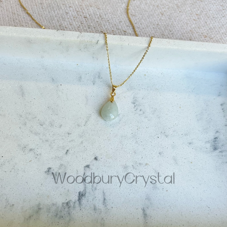 Puede incluir: Un delicado collar de cadena dorada con un colgante en forma de l&aacute;grima de color verde claro. El colgante est&aacute; hecho de una piedra natural y cuelga de una simple cadena dorada. El collar se muestra sobre una superficie de m&aacute;rmol blanco con el texto "WoodburyCrystal" en negro.