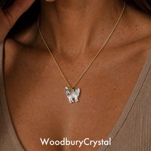 May include: A gold-colored necklace with a pendant featuring a white cat-shaped charm and a blue heart-shaped charm. The necklace is displayed on a person. The text "WoodburyCrystal" is at the bottom.