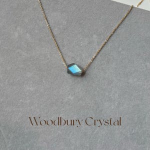 May include: A gold chain necklace with a single, faceted, iridescent blue-green gemstone pendant. The pendant is shaped like a diamond. The necklace is on a white background with the text "Woodbury Crystal" below the pendant.