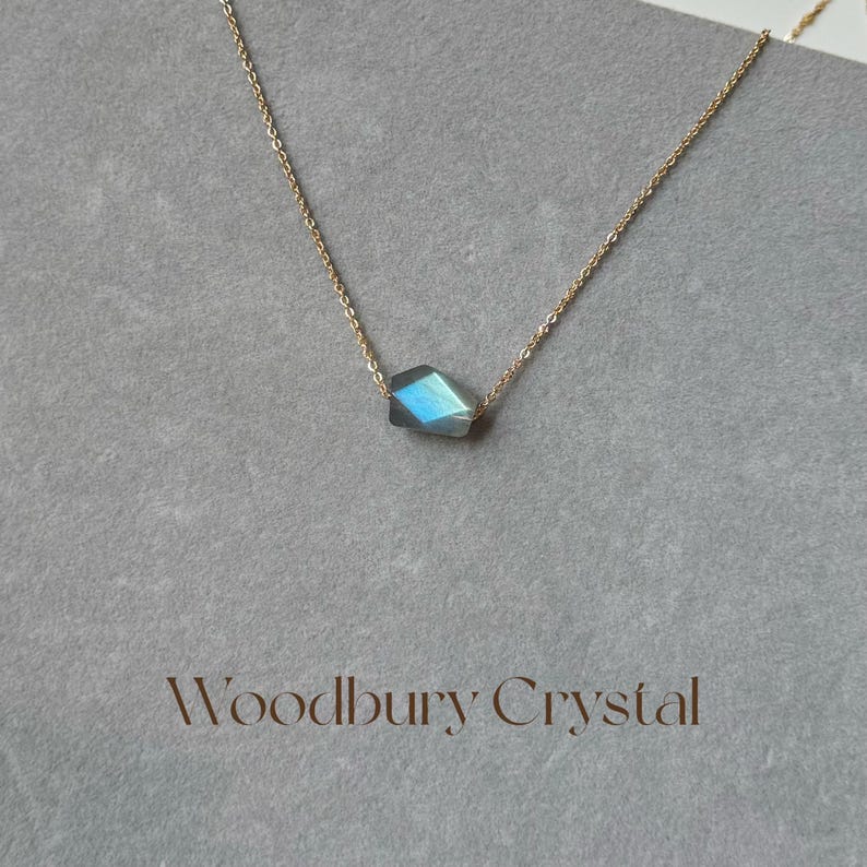 May include: A gold chain necklace with a single, faceted, iridescent blue-green gemstone pendant. The pendant is shaped like a diamond. The necklace is on a white background with the text "Woodbury Crystal" below the pendant.