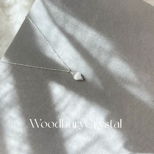 May include: A silver chain necklace with a white gemstone pendant. The pendant is round and has a smooth, polished surface. The necklace is on a gray background with the text "Woodbury Crystal" printed below the pendant.