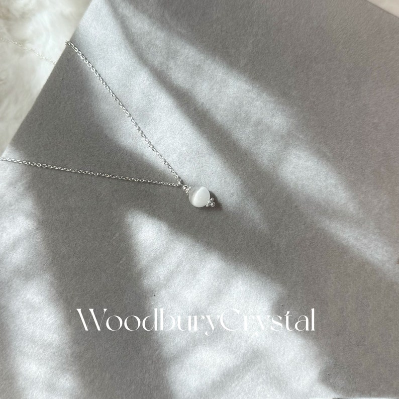May include: A silver chain necklace with a white gemstone pendant. The pendant is round and has a smooth, polished surface. The necklace is on a gray background with the text "Woodbury Crystal" printed below the pendant.