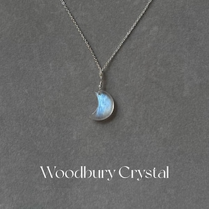 May include: A silver chain necklace with a crescent moon pendant made of iridescent blue moonstone. The pendant is attached to the chain with a silver bail. The necklace is on a gray background.  Woodbury Crystal.