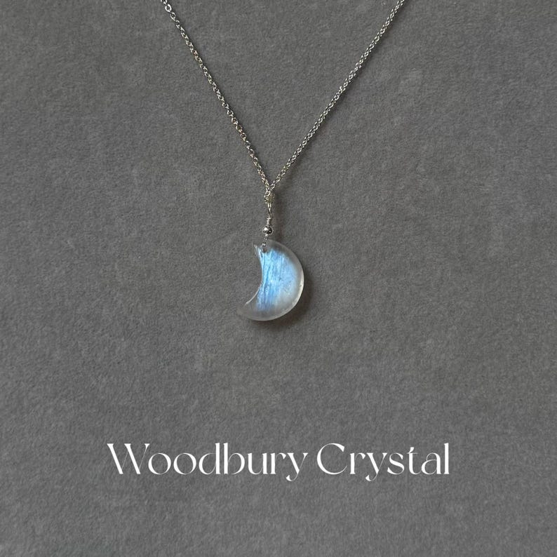 May include: A silver chain necklace with a crescent moon pendant made of iridescent blue moonstone. The pendant is attached to the chain with a silver bail. The necklace is on a gray background.  Woodbury Crystal.