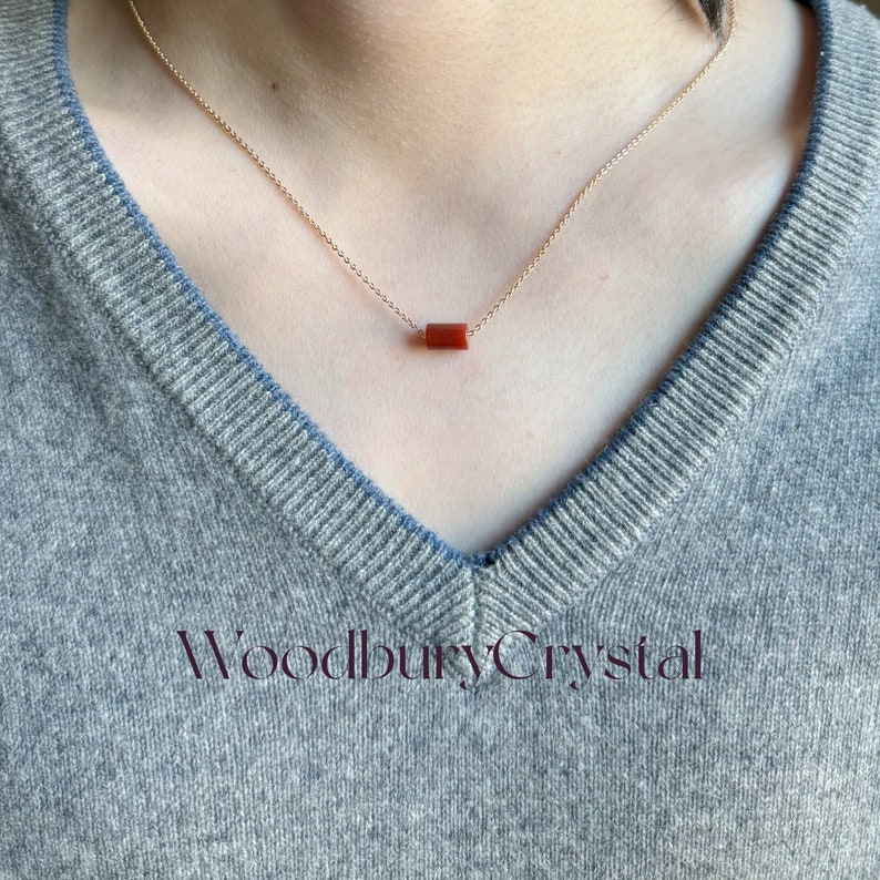 May include: A delicate gold chain necklace with a single, rectangular, red carnelian gemstone pendant. The necklace is worn by a person with a grey sweater.