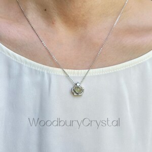 Puede incluir: Un collar de cadena de plata con un peque&ntilde;o colgante que presenta un cristal transparente en un engaste de corona. El colgante est&aacute; rodeado de cristales transparentes m&aacute;s peque&ntilde;os. El collar lo lleva una persona con una camisa blanca.