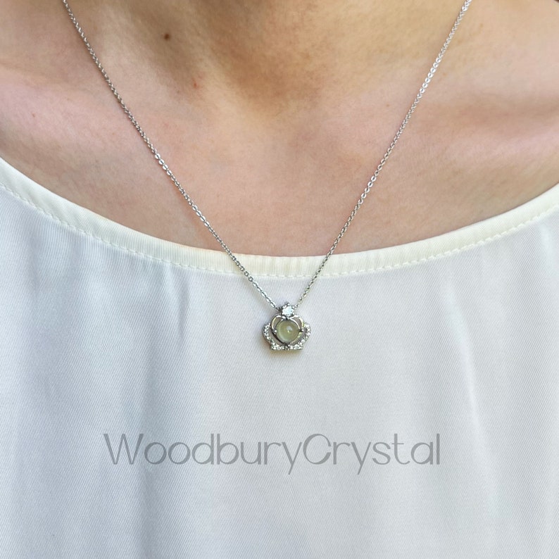 Puede incluir: Un collar de cadena de plata con un peque&ntilde;o colgante que presenta un cristal transparente en un engaste de corona. El colgante est&aacute; rodeado de cristales transparentes m&aacute;s peque&ntilde;os. El collar lo lleva una persona con una camisa blanca.