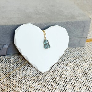 May include: A gold chain necklace with a blue-green labradorite pendant in the shape of a small, stylized human figure. The necklace is displayed on a white heart-shaped surface.