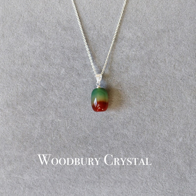 May include: A silver chain necklace with a green, red, and brown agate pendant. The pendant is oval shaped and has a smooth, polished finish. The necklace is on a gray background with the text "WOODBURY CRYSTAL" below the pendant.